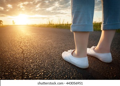 Woman in white sneakers standing on asphalt road towards sun. Concept of new start, travel, freedom etc.