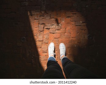 Woman In White Sneakers Stands In Sun Beam On Old Pavement Made Of Red Bricks. Modern Fashion. Light And Shadow.