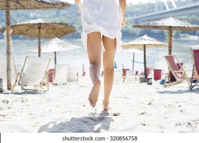 Woman In White Summer Dress Running On Sandy Beach Among Deck Chairs And Parasols, Rear View.