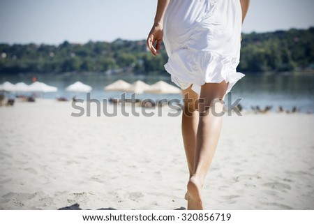 Woman in white summer dress running on sandy beach at river coast at summer burnout, rear view.