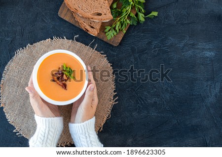A woman in a white sweater eats pumpkin soup with fried bacon with bread. Pumpkin cream soup in a white plate on a napkin on a black background. Top view. Copy space