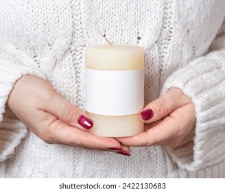 Woman in white sweater holding in hands cream candle with blank label, mock up, copy space, front view. Girl with red nails and pillar candle. Cozy holidays atmosphere - Powered by Shutterstock