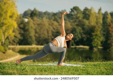 A woman in a white tank top and gray leggings performs a yoga pose on a mat in a park. The woman is smiling, looking upward and stretching her arms and legs. - Powered by Shutterstock