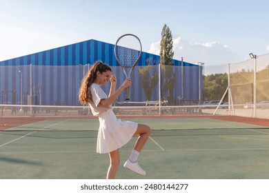 woman in a white tennis outfit celebrates a win with her racket raised high on a green tennis court - Powered by Shutterstock