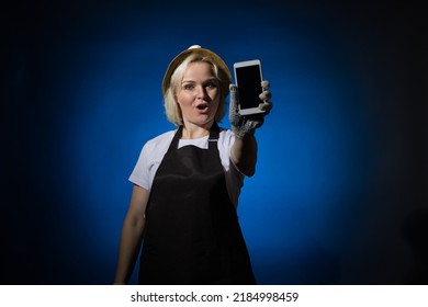 A woman in a white t-shirt, black apron, gloves and hat smiles and shows a phone with a blank screen on a dark background. - Shutterstock ID 2184998459