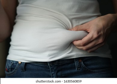 A woman in a white T-shirt and navy blue jeans squeezes a fold of fat on her belly, closeup, selective Focus.