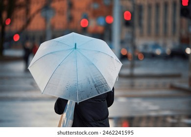 Woman with white umbrella waiting signal for cross walk during rain in the evening. Alone woman stands with white umbrella, back view. City rain scene
