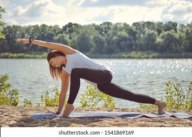 Woman In White Undershirt And Black Leggings Is Practicing Yoga Performing Yoga-asanas On A Mat Outdoors, Near A Riverside.