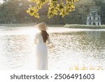 Woman in white Vietnamese traditional dress and conical hat at Hoan Kiem Lake in Hanoi, Vietnam. Summer vacation and holidays. Smiling Asian women relaxing in outdoors. Travel and lifestyle Concept.