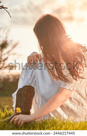 A woman in a white, wavy dress and cowboy boots picking a dandelion in a meadow with the sunset behind her. 
