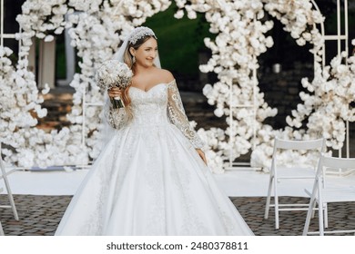 A woman in a white wedding dress is standing in front of a white archway. She is holding a bouquet of flowers and she is posing for a picture. Concept of elegance and celebration - Powered by Shutterstock