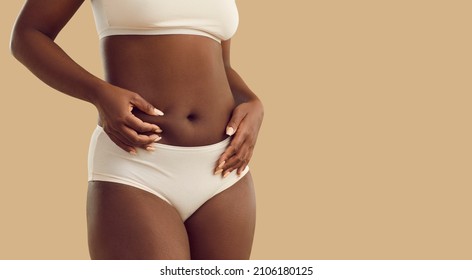 Woman who has larger size and type of figure pinches her skin standing on beige background. Cropped closeup image of belly of unknown black leather woman in white cotton lingerie. Banner. Copy space.