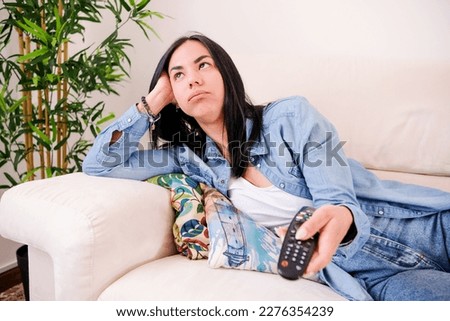 Woman who is lying on her sofa, feeling bored and uninterested while zapping TV channels with her remote control