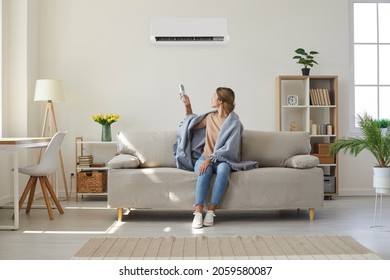 Woman who's sitting on sofa under warm plaid in living room switches off her air conditioner on wall. Young girl adjusting modern AC system, regulating temperature and enjoying cool fresh air at home