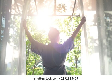 The woman was at the window in the bedroom. She opened the curtains on the window. In the morning and she looks at the view of mountains and trees at sunrise.