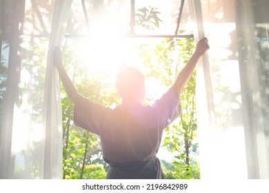The woman was at the window in the bedroom. She opened the curtains on the window. In the morning and she looks at the view of mountains and trees at sunrise. - Shutterstock ID 2196842989