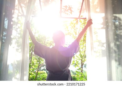 The woman was at the window in the bedroom. She opened the curtains on the window. In the morning and she looks at the view of mountains and trees at sunrise. - Shutterstock ID 2210171225