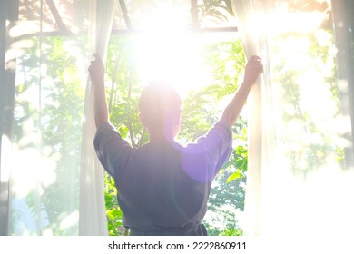 The woman was at the window in the bedroom. She opened the curtains on the window. In the morning and she looks at the view of mountains and trees at sunrise. - Shutterstock ID 2222860911