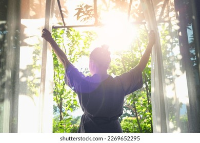 The woman was at the window in the bedroom. She opened the curtains on the window. In the morning and she looks at the view of mountains and trees at sunrise. - Shutterstock ID 2269652295
