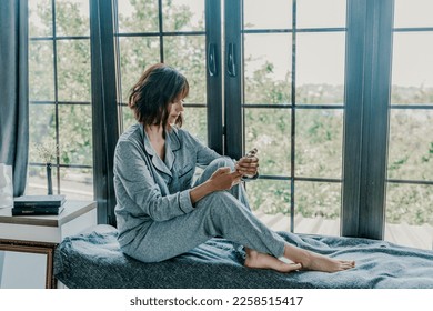 Woman window sill pajamas. Resting woman in pajamas. A calm girl with a phone sits on the windowsill at home. Side view - Shutterstock ID 2258515417