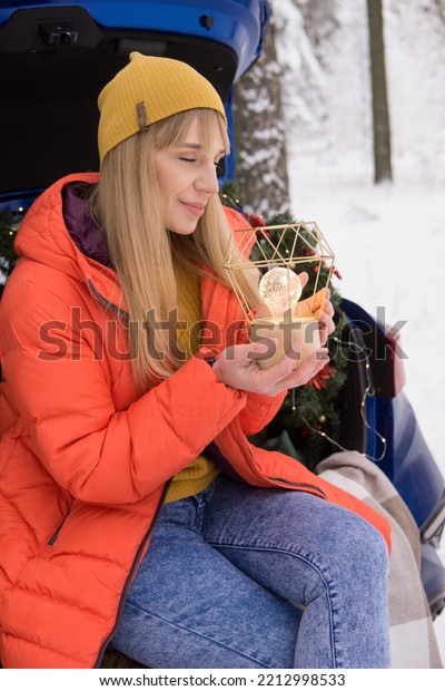 A woman in a winter snow-covered forest in the\
trunk of a blue car decorated with Christmas decor. The concept of\
Christmas and winter\
holidays.