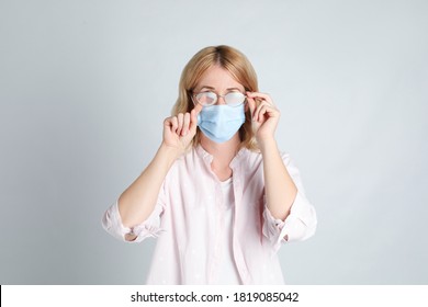 Woman Wiping Foggy Glasses Caused By Wearing Medical Mask On Light Background