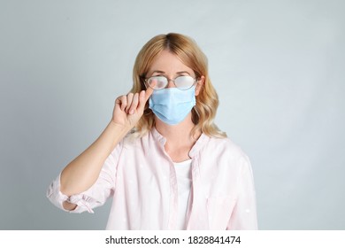 Woman Wiping Foggy Glasses Caused By Wearing Medical Mask On Light Background