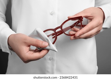Woman wiping her glasses with microfiber cloth on grey background, closeup