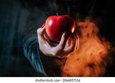 Woman as witch in black offers red apple as symbol of temptation, poison. Fairy tale, white snow wizard concept. Spooky halloween, cosplay. Smoke, haze background.