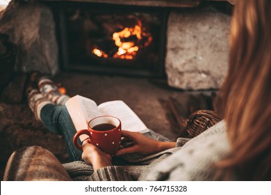 Woman in woollen socks by the fireplace. Unrecognisable relaxes by warm fire with a cup of hot drink and warming up her feet in woollen socks. Cozy atmosphere. Winter and Christmas holidays concept.