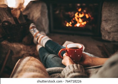 Woman In Woollen Socks By The Fireplace. Unrecognisable Relaxes By Warm Fire With A Cup Of Hot Drink And Warming Up Her Feet In Woollen Socks. Cozy Atmosphere. Winter And Christmas Holidays Concept.