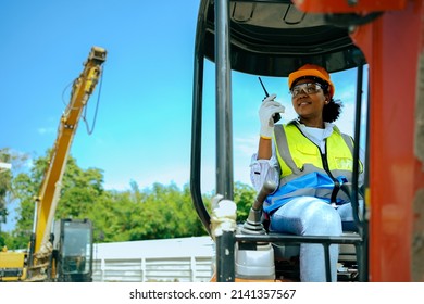 A woman worker driving a backhoe to dig a hole in a construction site holding a walkie talkie.African American female engineer wearing a hard hat and vest.
				cute female with black skin gender equality.