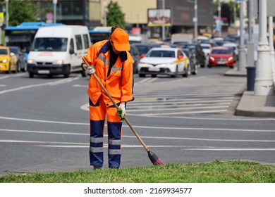 Woman Worker In Orange Uniform With A Broom Sweeps The Road On Cars Background. Street Cleaning In Summer City