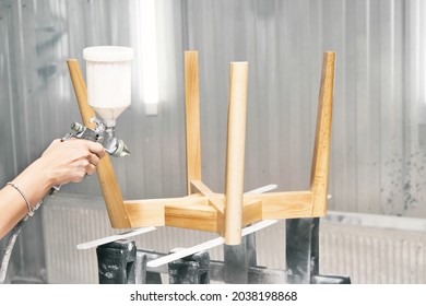 Woman Worker Paints Wooden Chair Base In Transparent Varnish With Spray Gun On Rack Support In Contemporary Spraying Booth Close View