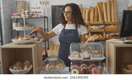 Woman working behind the counter of a bakery shop with various pastries and breads on display, wearing an apron and glasses, selecting desserts with tongs in a cozy interior setting. - Powered by Shutterstock