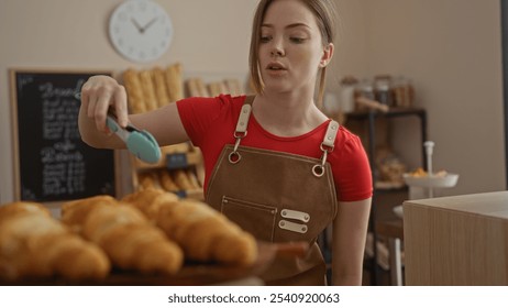 Woman working in a cozy bakery shop wearing a red shirt and apron, arranging freshly baked croissants on a tray - Powered by Shutterstock