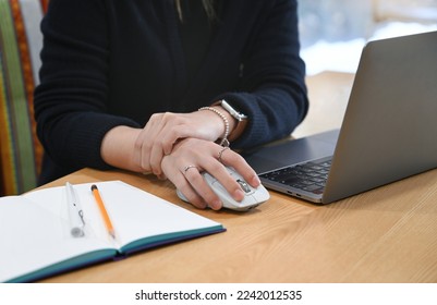 A woman working at a desk using a laptop and mouse is complaining of wrist joint pain - Shutterstock ID 2242012535