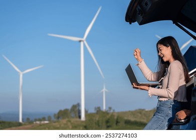 A woman working with her car trunk open and gazing at her laptop, content with a job well done in the midst of a wind farm that generates renewable energy - Powered by Shutterstock