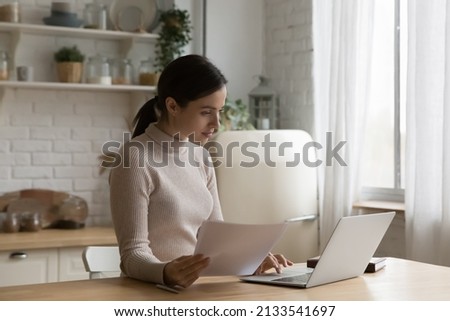 Woman working from home seated at table in domestic kitchen use laptop, prepare official papers documents for client, holds sheet comparing information, busy in paperwork, pay bills via e-bank concept