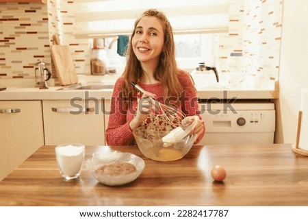 The woman working in the kitchen puts the ingredients in the bowl in front of her to make cookies. Image of woman working in kitchen in warm tones.