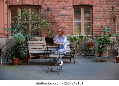 Woman working laptop in a cozy courtyard with potted plants. Concept of remote work or freelancing