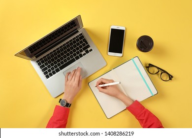 Woman Working With Modern Laptop And Writing In Notebook At Color Table, Top View