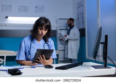 Woman Working As Nurse With Digital Tablet For Checkup At Night. Medical Assistant Using Modern Device And Computer On Desk For Healthcare And Treatment, Working Late. Person With Job
