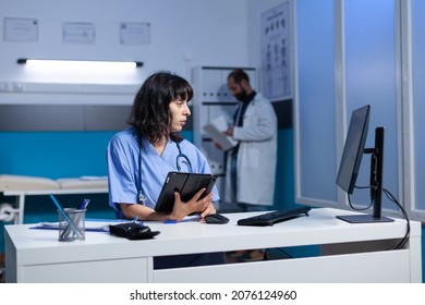 Woman Working As Nurse With Tablet For Checkup At Night. Medical Assistant Looking At Modern Device And Computer On Desk For Healthcare And Treatment, Working Late. Person With Job