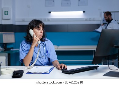 Woman Working As Nurse Using Telephone For Conversation With Patient About Healthcare Appointment. Medical Assistant Talking On Landline Phone For Remote Communication, Working Late.