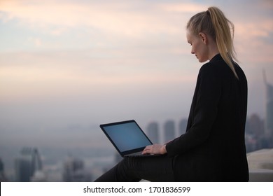 Woman working on computer & sitting at the edge of skyscraper rooftop during sunrise. Beautiful city background view.