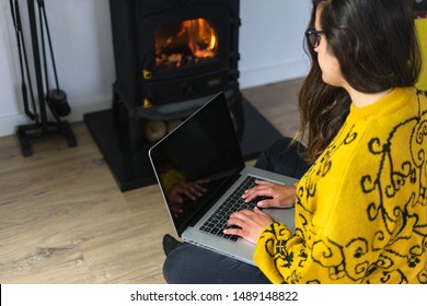 Woman Working On Her Laptop Computer In Front Of A Wood Burning Stove Or Fireplace.