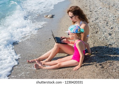 Woman Working On A Laptop At The Beach, Copy Space. Family Vacation, Summer Vacation. Mom And Daughter Are Sitting On The Beach With A Laptop, Distant Work.