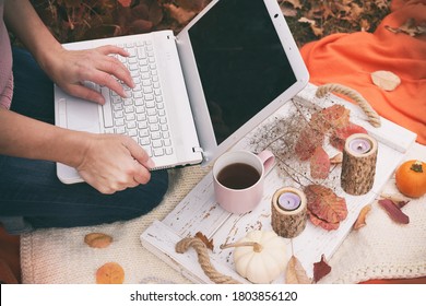 Woman working on laptop in the garden. Next to it is a tray with a cup of tea and autumn decorations. Remote work. Quarantine work at home.
