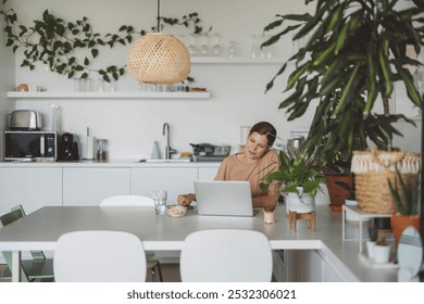 A woman working on a laptop at a modern kitchen table, working from home, surrounded by plants. The kitchen features white cabinetry, a pendant light, and a cozy atmosphere. - Powered by Shutterstock